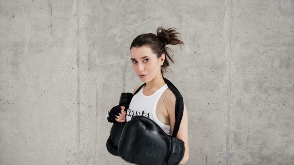 Confident young woman posing with boxing gloves against a concrete wall, showcasing style and sportiness.